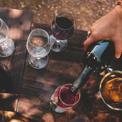 Person pouring red wine into a glass on a wooden table with other wine glasses.